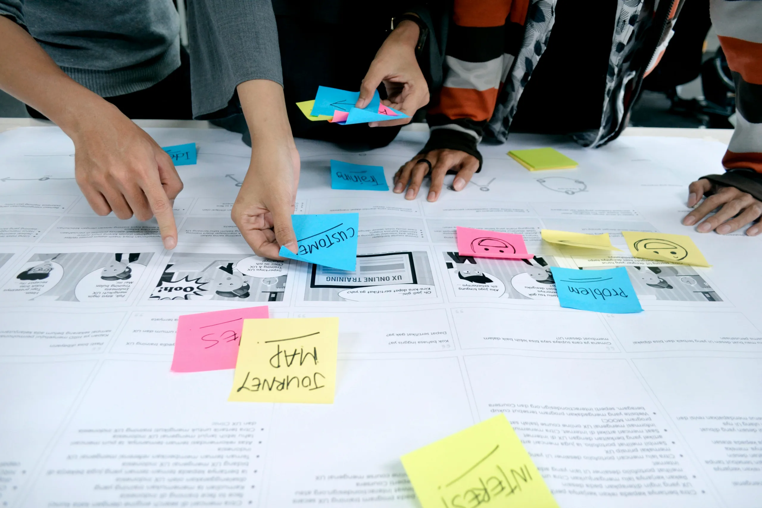 Several people collaborate around a table covered with printed layouts and colorful sticky notes, pointing and organizing ideas together.