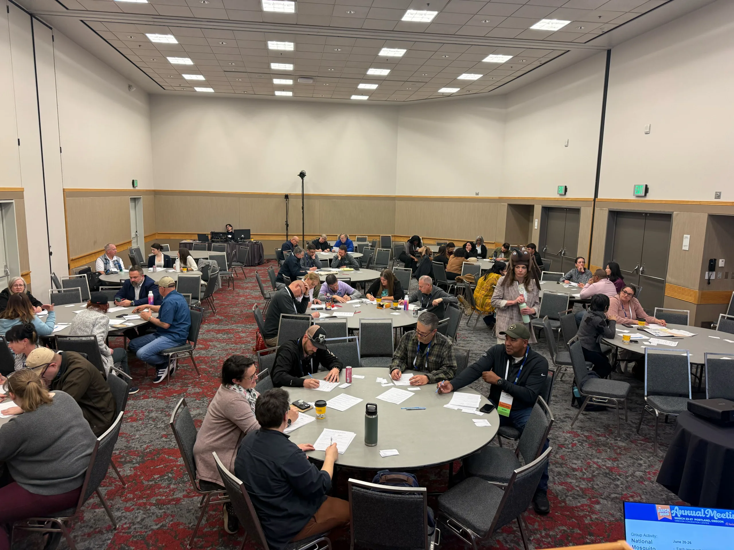 A large conference room with high ceilings and bright overhead lighting, where multiple small groups of adults sit around round tables.