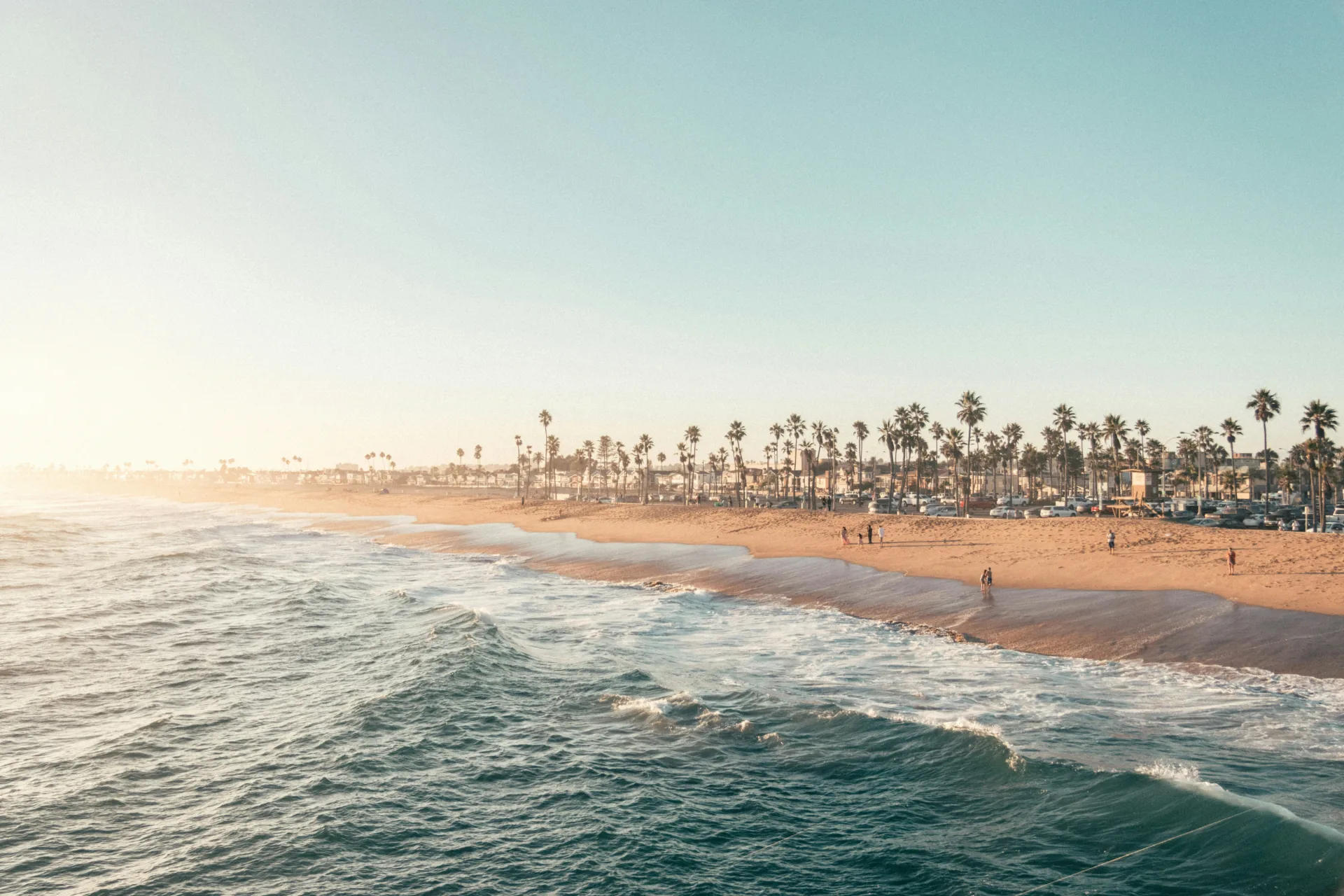 A wide, sunlit coastal scene showing gentle ocean waves rolling onto a sandy beach. A line of tall palm trees and a busy beachfront with parked cars and scattered people stretches along the shore in the background under a clear, hazy sky.
