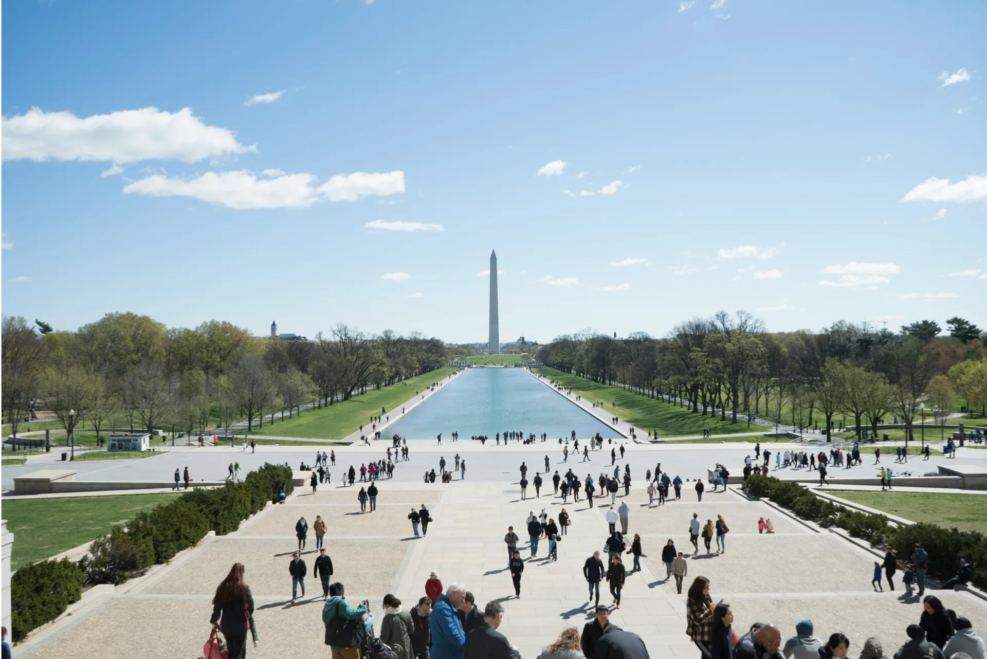 People gather on the steps of the Lincoln Memorial overlooking the Reflecting Pool, with the Washington Monument centered in the distance beneath a bright blue sky.